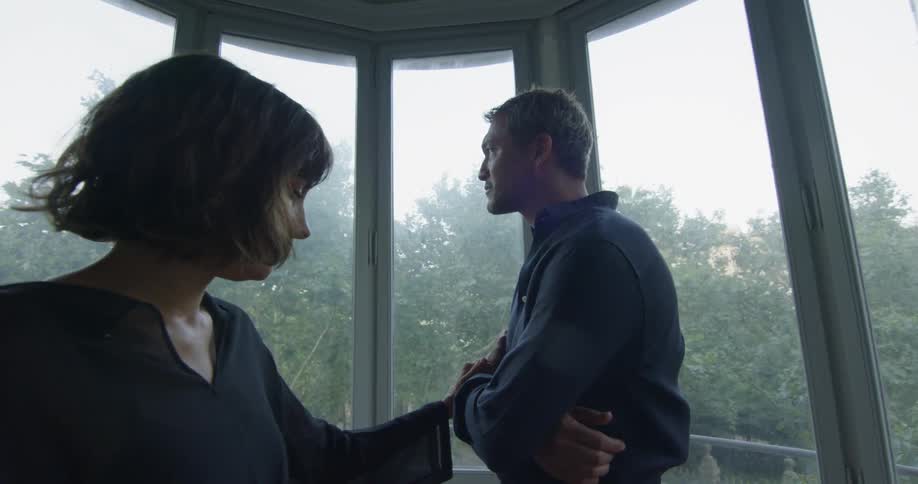 A brunette woman with chin length hair comforts a man with his arms crossed next to a set of wall length windows