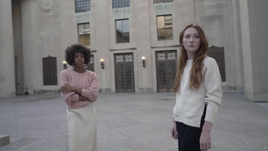 A woman with long red hair draped over her shoulders stands with a black woman who is posing confidently in a building with many columns and a statue