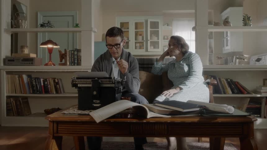 A bespectacled man is sitting on a sofa and is intensely focused on what he's writing on the typewriter then his wife enters the room to place a cup of coffee in front of him