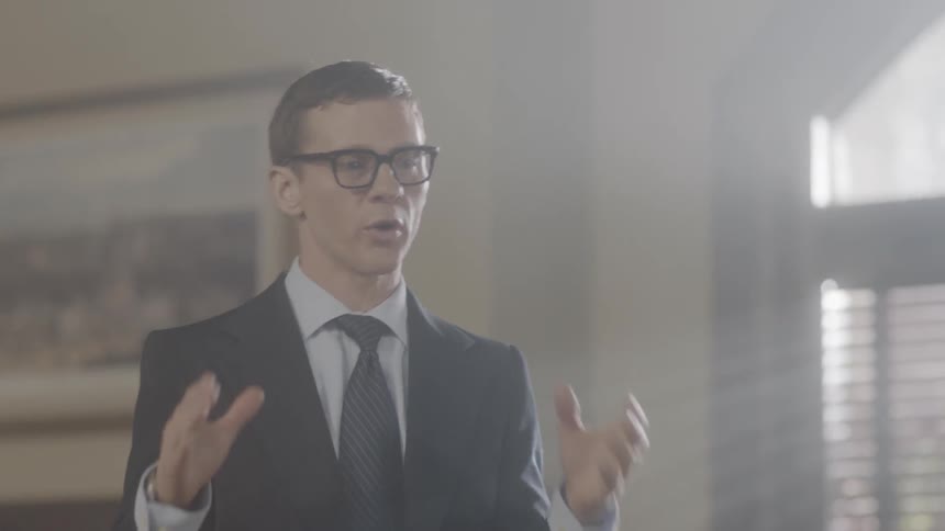 A man talks and gestures near a presentation in front of a group of people