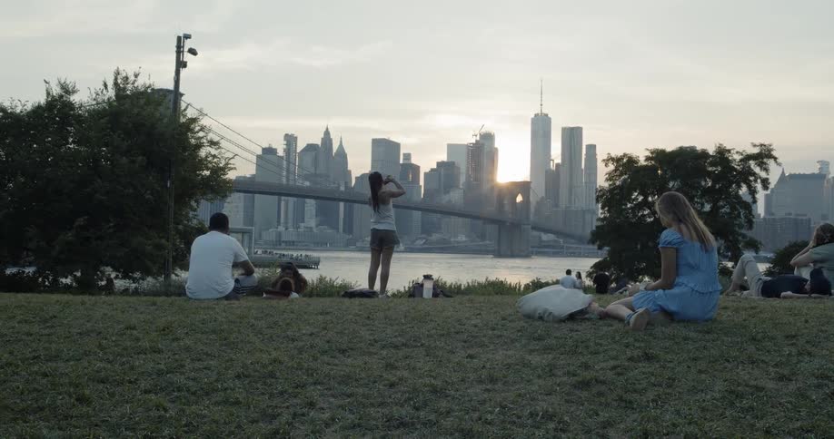Several people are relaxing in a park that is across a body of water from a dense metropolitan area