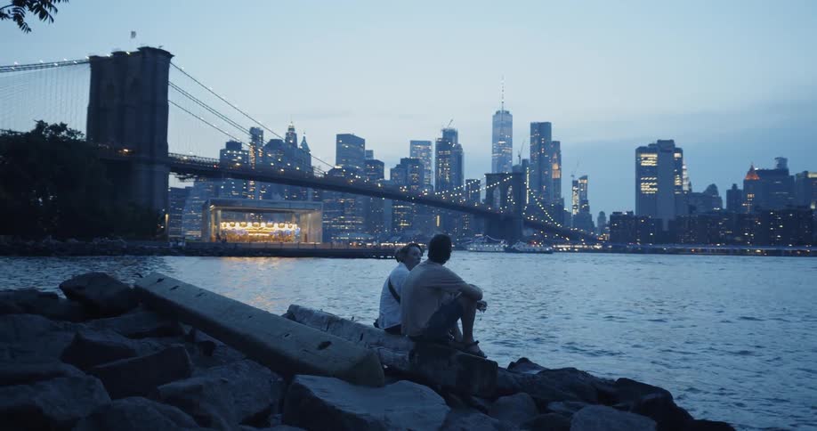 A man and woman sitting together at a Brooklyn riverside in the evening