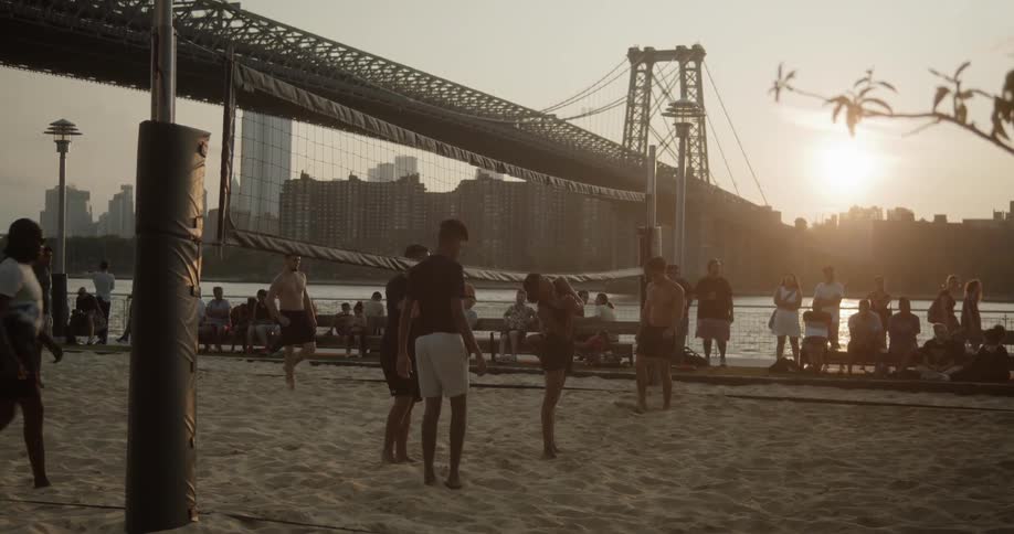 Young men and women play volleyball on the sand in a park while others sit on benches and watch