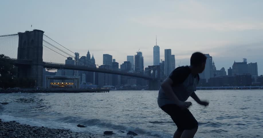 A boy skipping rocks across a river facing the Brooklyn skyline at dusk