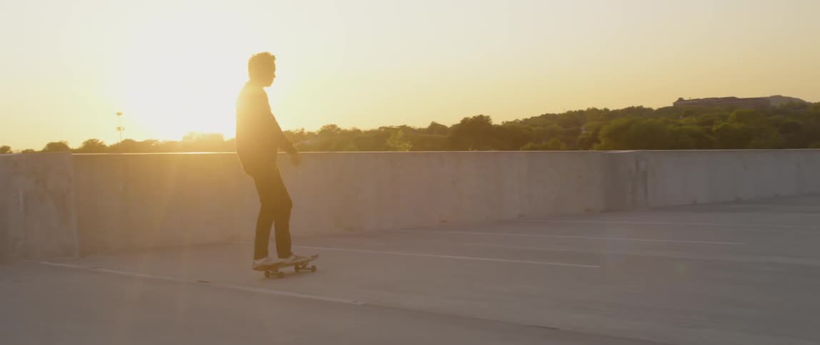 A man slips off his skateboard while riding on a rooftop parking area at sunset
