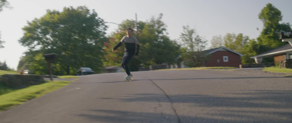 A young man skateboards down a street in a suburb and eventually falls off the board