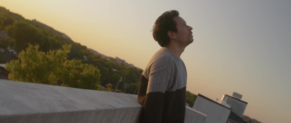 A man in a sweater stands on a rooftop at sunset