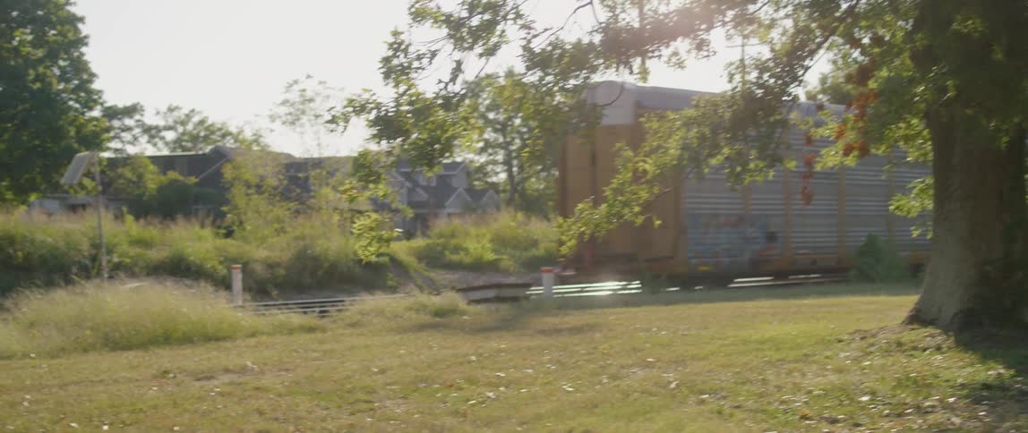 The last box car of a train slowly passes by on the tracks next to a grassy area with trees and bushes
