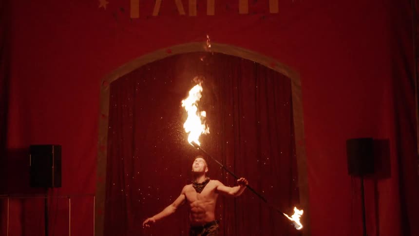 A fire eater holding a flaming baton breathes out a tall flame in front of an audience