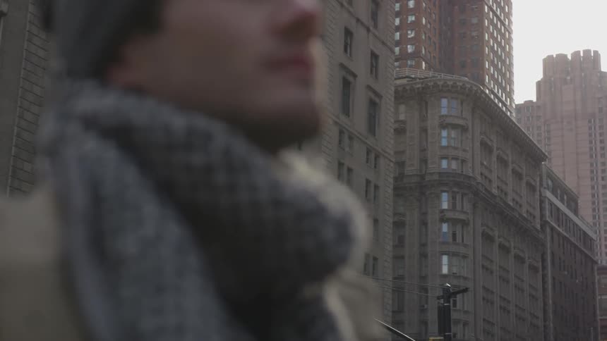 A man wearing a hat and scarf stands on a city sidewalk and looks up at the buildings