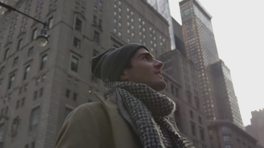 A man in a scarf and coat stands on a city sidewalk surrounded by skyscrapers