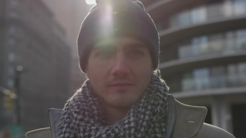 A man stands on a city sidewalk staring into the camera while traffic moves behind him