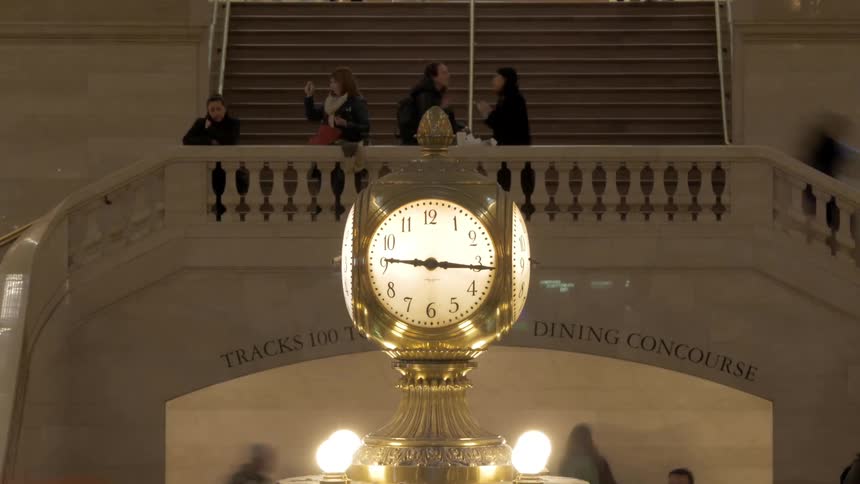 People walk up and down the steps and stand at the railing of grand central station overlooking the clock