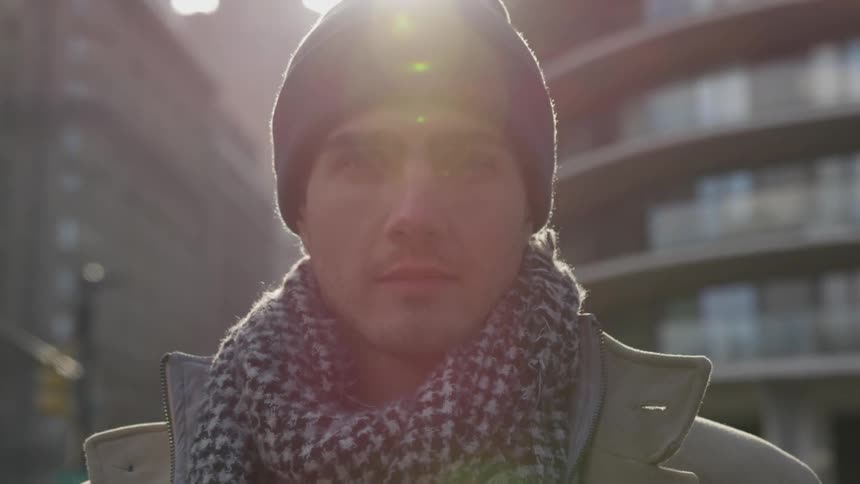 Man in a scarf and hat stares intently into the camera with a city behind him