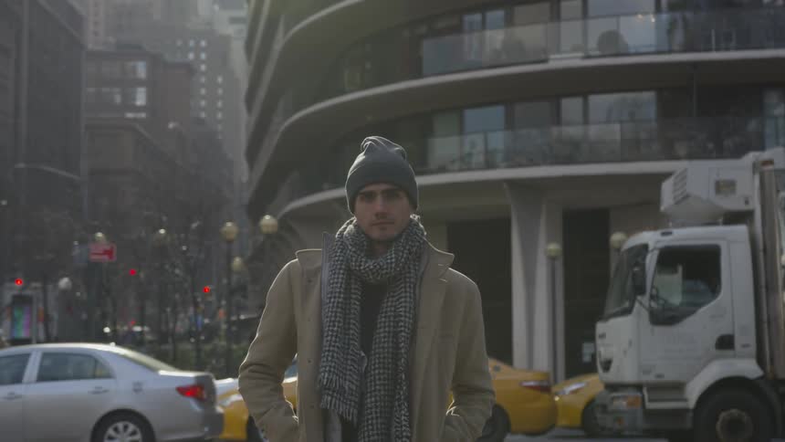 A man with a coat and scarf standing on a city street with cars in the background