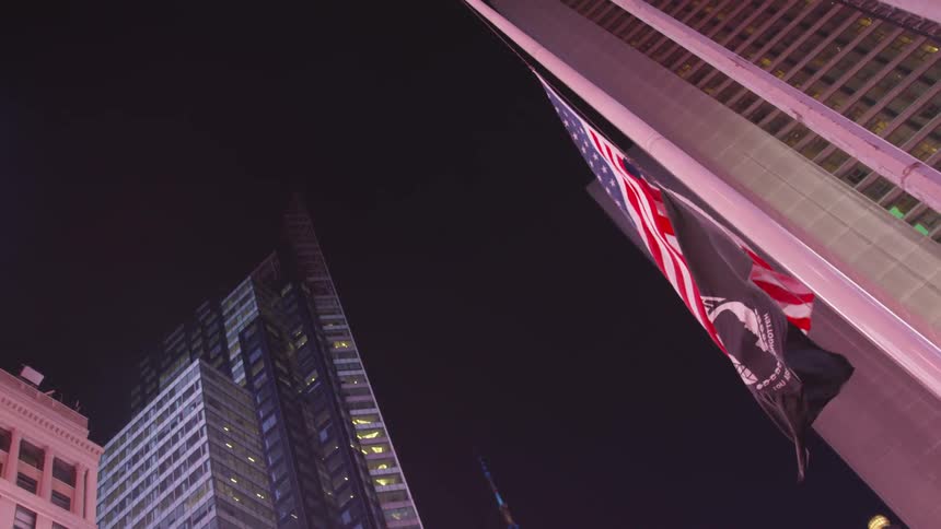 An american flag and a pow flag flapping in the wind in the city at nighttime