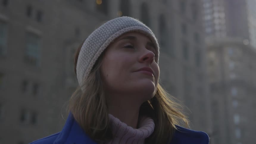 A woman in a headband and coat stands on a city sidewalk near skyscrapers