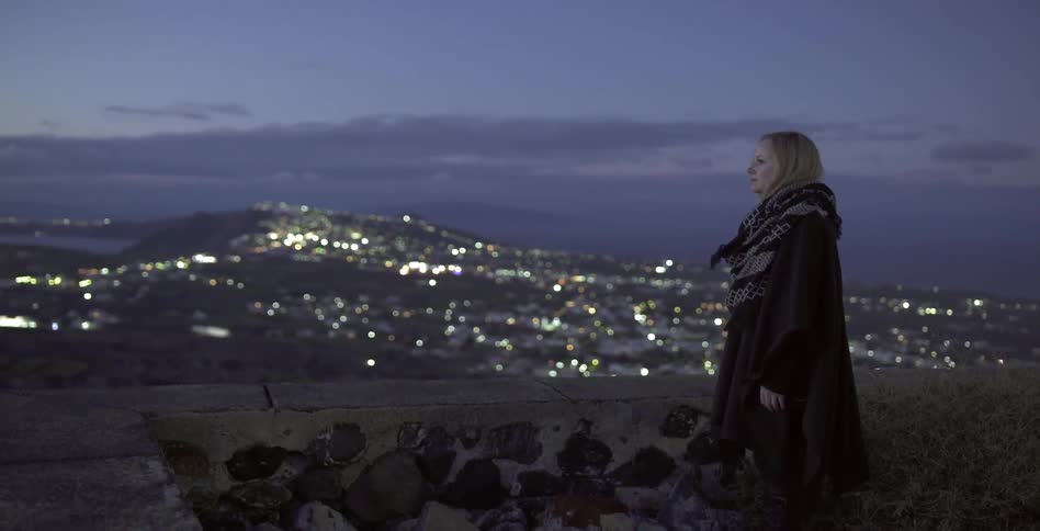 A woman overlooks a city at night from a hilltop