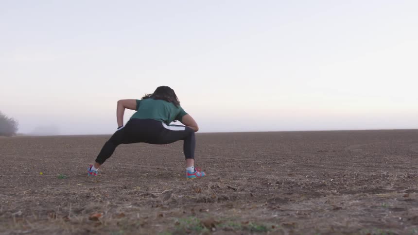 A young woman wearing exercise pants and a green t shirt stretches her legs in a field