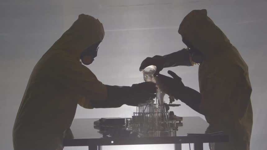 Two people in yellow hazardous materials protective suits stand across from each other over a single table and assist each other in pouring bubbling liquids from one glass container to another