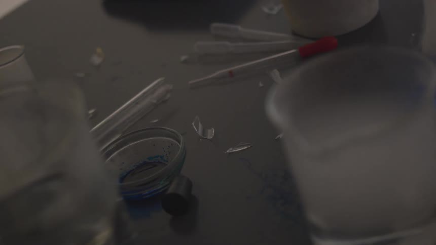 Droppers and laboratory beakers sit on a table as a person walks near by