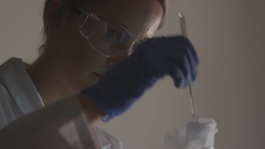 A woman with goggles on her face wearing a white lab coat stirs a smoking beaker with a blue gloved hand