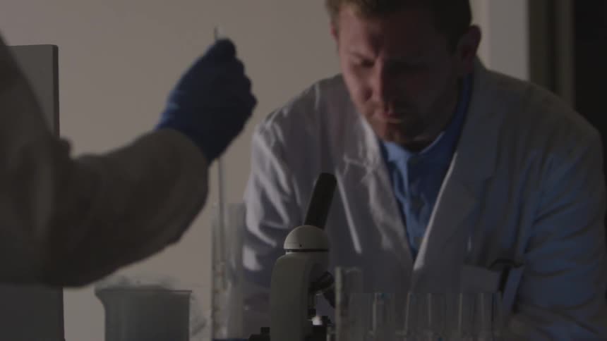 Two scientists work together in a lab looking through a microscope and putting a glass stirrer in a beaker