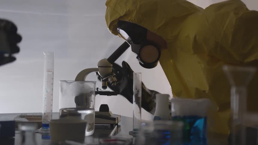 A person in head to toe yellow hazardous materials protective suit is also wearing goggles and a respirator as they look into an outdated microscope that is located on a table amidst several pieces of glassware with bubbling colored liquids in them
