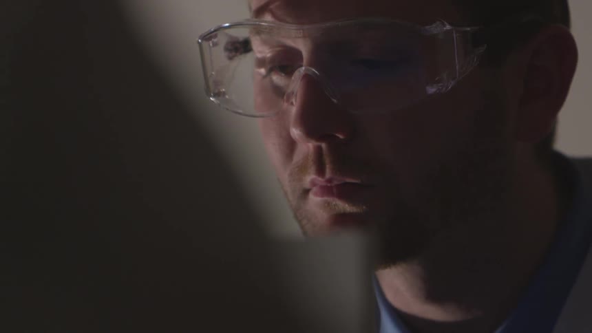 A white man wearing safety glasses looks at a computer monitor