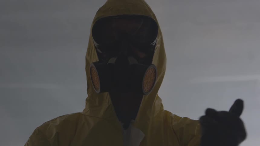 A person in a yellow containment suit and wearing a gas mask pulls on a pair of black rubber gloves