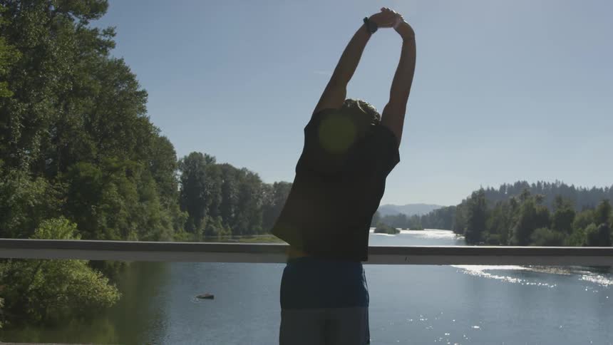 A person standing on a bridge stretches while looking at the water