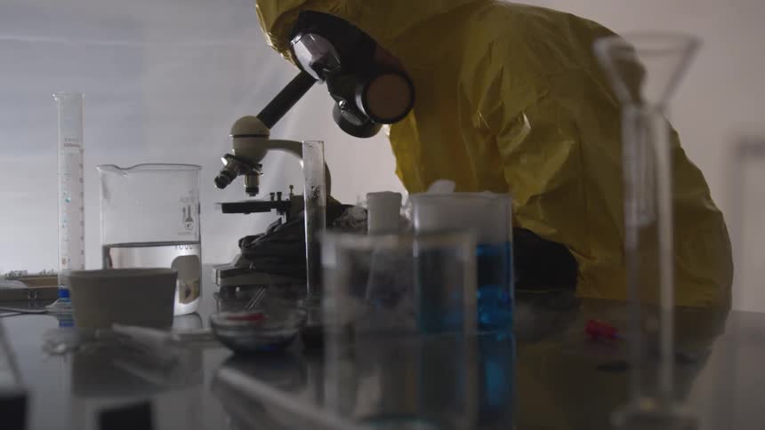 A person in a yellow lab suit and wearing a gas mask peers through a microscope in a laboratory