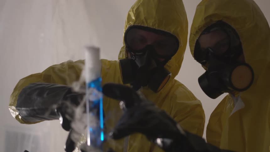 Two people in yellow hazardous material protective suits complete with gloves goggles and respirators inspect a glass graduated cylinder that is full of a bubbling blue liquid that is producing smoke