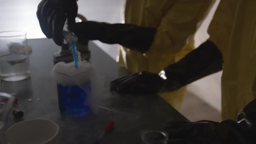 Two lab workers in safety gear and rubber gloves pour a blue smoking liquid into a glass beaker