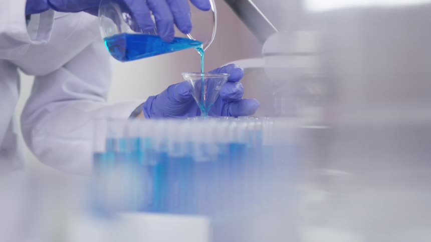 A person in a white lab coat pouring a blue chemical into a funnel