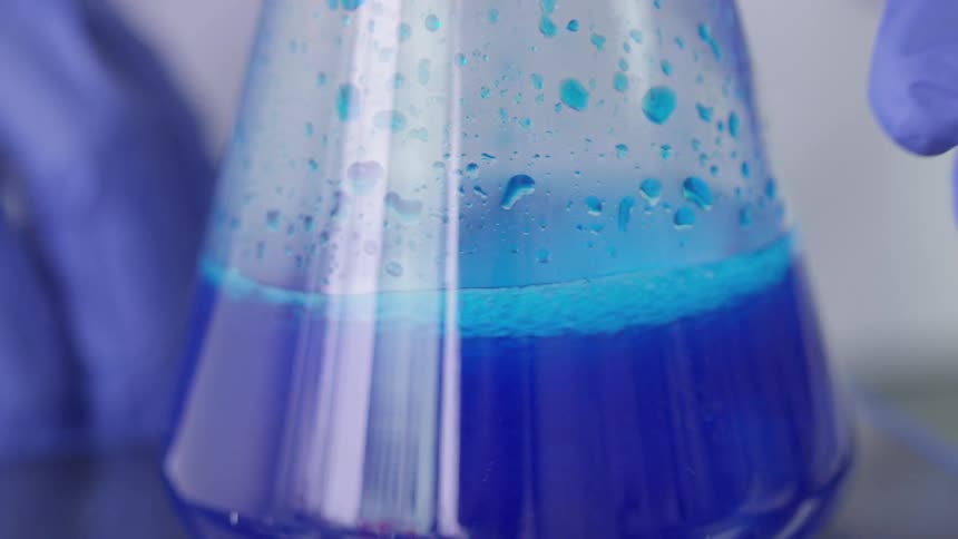 A blue liquid swirls around a container and is then picked up by a glove wearing hand