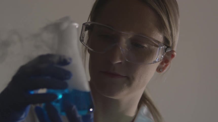A young woman wearing goggles gloves and a lab coat holds a beaker full of bubbling steaming blue liquid