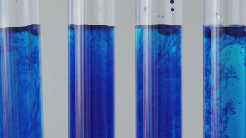 Each of four test tubes of blue liquid have a darker blue liquid dripped into them by a glass pipette and the liquids can be seen mixing and blending together