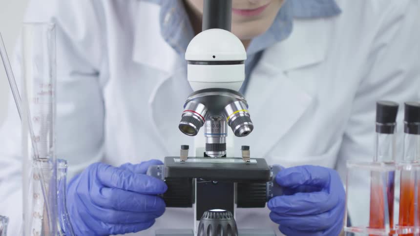 A lab with blue gloves technician is slowly turning the knobs on a microscope with test tubes beside it