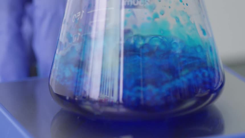 An erlenmeyer flask containing a magnetic stir bar has a blue liquid added to it and the magnetic stirrer beneath it is turned of an begins to quickly stir the liquid