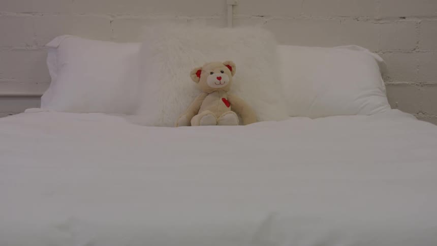 A brown teddy bear with a red heart on a white bed with pillows
