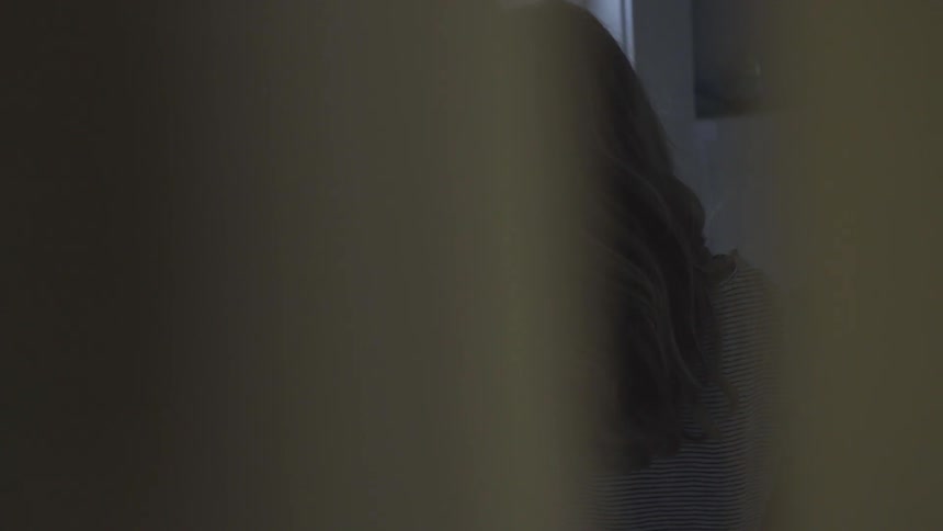 A female with long brown hair can be see standing before bathroom sink through a partially opened door