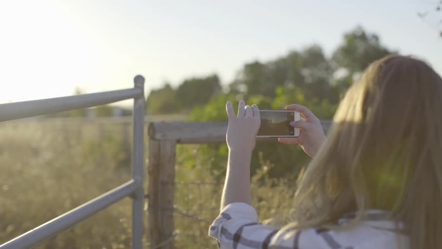 A woman takes a photo of a field that's beyond a fence
