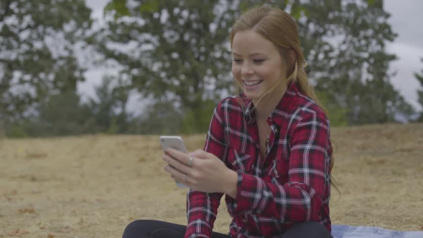 A woman sits on a blanket in a field and talks on her phone as she smiles