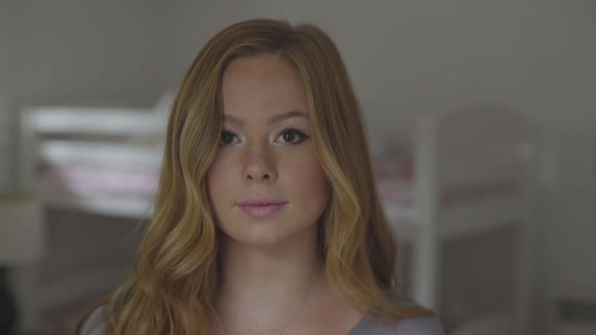 A woman stares into the camera while standing in a bedroom