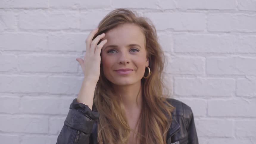 A woman runs her hand through her hair and smiles as she stands in front of a wall
