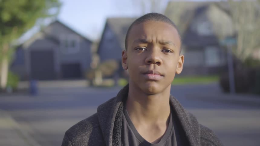 A teenage boy stares into the camera while standing on a neighborhood sidewalk