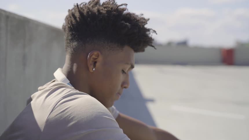 A young black man sits and looks down at the ground while rubbing his head