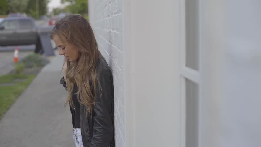 A young woman in a leather jacket stands on a sidewalk with her back against the side building as she brushes her hair from her face while looking at the ground