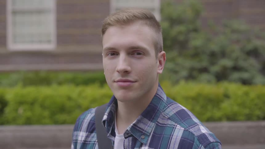 A young man in front of a brick building and bushes turns to the camera and smiles slightly